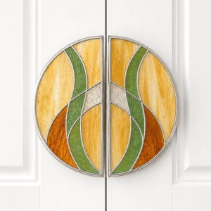 Puede incluir: Un par de tiradores decorativos para armarios. Cada tirador es un semicírculo con un diseño de vidriera. El vidrio presenta formas curvas amarillas, verdes y marrones, enmarcadas por un borde de metal plateado. Los tiradores están montados en una puerta de armario blanca.