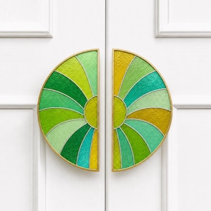 Puede incluir: Dos manijas de puerta semicirculares con un diseño de vidriera. Las manijas presentan un marco de metal dorado y un colorido patrón de vidrio verde, amarillo y turquesa. Están montadas en una puerta blanca.