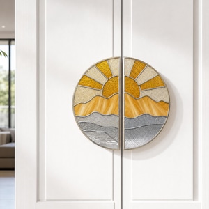 Puede incluir: Tiradores de puerta decorativos con diseño de vidriera. Los tiradores redondos representan un amanecer sobre una cordillera. El sol es amarillo y dorado, las montañas son amarillas y beige, y el agua es gris. Montados en una puerta blanca.