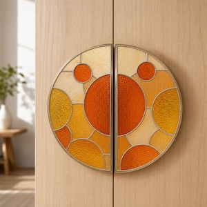 May include: A pair of semi-circular cabinet handles featuring a stained glass design with various circular shapes in shades of orange, yellow, and cream, encased in a thin brass-toned metal frame, mounted on a light-coloured wooden cabinet door.