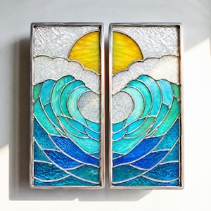 May include: Two rectangular stained glass panels featuring a sun and ocean wave design. The panels display a yellow sun against a textured white sky, with layered blue and teal glass waves in the foreground, all encased in a silver metal frame.