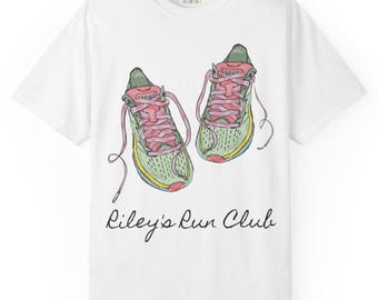 Puede incluir: Camiseta blanca con una ilustración en acuarela de zapatillas para correr. Las zapatillas son de color verde claro con cordones rosas y detalles amarillos. El texto "Riley's Run Club" está escrito en cursiva debajo.