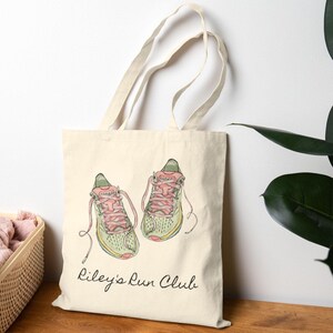May include: Cream-colored tote bag featuring a watercolor illustration of running shoes with pink laces and green accents. The text "Riley's Run Club" is written in a cursive font below the shoes. The bag has a natural canvas texture.