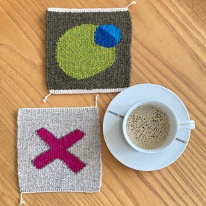 Op de afbeelding: Twee geweven onderzetters, een met een groene cirkel met een blauwe cirkel erin, en de andere met een roze X op een beige achtergrond.