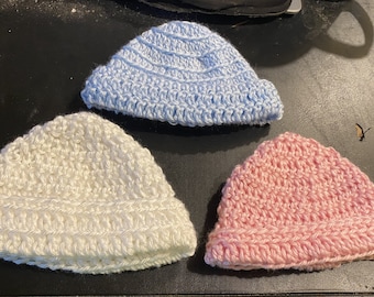 Könnte beinhalten: Drei gehäkelte Mützen in verschiedenen Farben: hellblau, weiß und rosa. Die Mützen haben alle den gleichen Stil mit einer einfachen, abgerundeten Form.