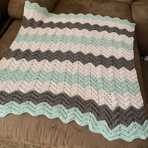 Può includere: Una copertina per neonati all'uncinetto con un motivo a chevron in tonalità di grigio, bianco e verde chiaro.