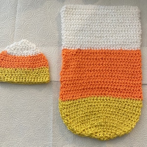 May include: A crocheted candy corn hat and a matching crocheted candy corn baby blanket. The hat is white, orange, and yellow. The blanket is white, orange, and yellow.