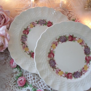 May include: Two white ceramic plates with a floral design. The plates have a raised, woven pattern and are decorated with a band of pink, red, and purple flowers.