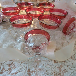May include: Set of clear glass teacups with a ruby red rim and handle. The cups have a decorative pattern and are arranged on a lace tablecloth. A pale pink ribbon is draped across the cups.