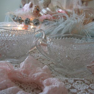 May include: Two clear glass cream and sugar dishes with ornate, embossed patterns. The creamer has a spout and handle, while the sugar bowl has two handles. Both sit on a lace-covered surface, suggesting a vintage or antique aesthetic.