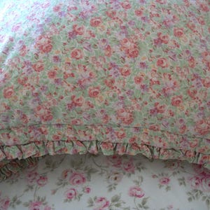 May include: A floral pillow with a ruffled edge. The pillow features a pattern of pink and red roses on a light green background. The pillow is part of a bedding set with a matching floral pattern.