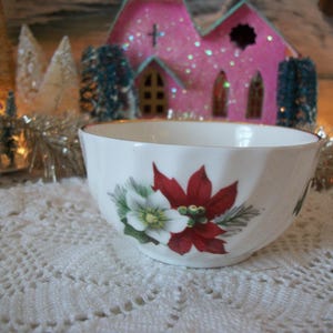 May include: White ceramic bowl with a gold rim, decorated with a poinsettia and greenery design. The bowl is set on a white lace tablecloth, with a pink church and decorative trees in the background.