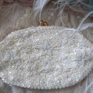 May include: An ivory-colored, oval-shaped beaded clutch. The clutch is covered in small, iridescent sequins and features star-shaped beaded designs. A gold-toned clasp secures the top. The background includes lace and feathers, suggesting a vintage or bridal theme.