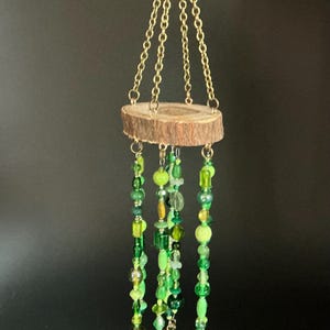 May include: A hanging mobile featuring a circular wooden slice with natural bark edges. Three gold-toned metal chains attach to the top. Multiple strands of green glass and plastic beads in various shapes and sizes hang from the wood, ending in small metal bells.