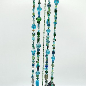 May include: A hanging mobile featuring a circular wooden slice top with silver chains. Multiple strands of blue and green glass beads in various shapes and sizes hang from the wood, ending with iridescent shell-like pieces at the bottom.