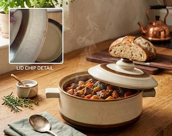 May include: A round ceramic casserole dish with a speckled cream and brown glaze, featuring two side handles and a matching lid. The dish contains a beef stew with visible carrots and potatoes. An inset shows a small chip on the lid edge.