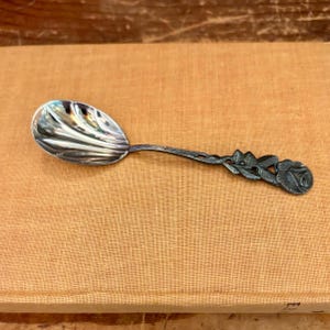 May include: A silver-toned metal spoon featuring a bowl with a shell-like fluted design. The handle is detailed with a sculpted, three-dimensional rose motif, including textured leaves and a blooming flower at the end of the stem.