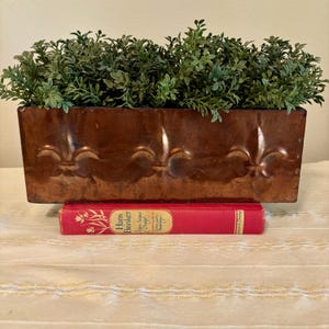 May include: A rectangular copper-toned metal planter featuring embossed fleur-de-lis patterns, filled with green artificial foliage. The planter rests on a red hardcover book titled Hans Brinker by Mary Mapes Dodge.