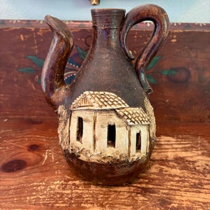 May include: A dark brown ceramic pitcher featuring a relief sculpture of a small house with a tiled roof and three open doorways. The vessel has a curved handle and a spout, with a textured finish and a contrasting light cream color on the house.