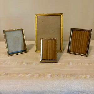 May include: A collection of four gold-toned metal picture frames. One large vertical frame stands in the back, flanked by two smaller frames on the sides, and one small vertical frame is positioned in the front center.