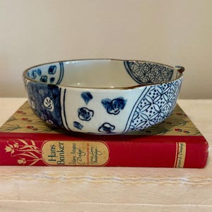 May include: A shallow ceramic bowl with a white interior and a blue and white patterned exterior featuring geometric and floral motifs, resting on a red hardcover book titled Hans Brinker by Mary Mapes Dodge.