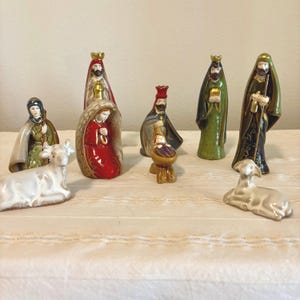 May include: A ceramic nativity set featuring nine hand-painted figurines including a central infant in a manger, kneeling figures in red and green robes, standing figures with gold crowns, and two small animal figures in white and gray tones.