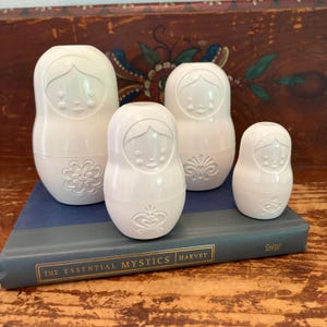 May include: A set of four white nesting dolls with embossed facial features and floral patterns on the lower sections. The dolls are arranged in a row on top of a dark blue book titled The Essential Mystics by Harvey, which is placed on a wooden surface.
