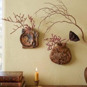 May include: Two brown ceramic wall-mounted vases with textured surfaces. The left vase holds small pink flowers, and the right vase holds pink flowers and a dark dried lotus pod. A yellow beeswax candle in a brass holder sits below.