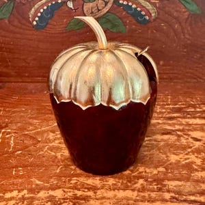 May include: A dark red, textured, strawberry-shaped container with a metallic gold-toned lid. The lid features a sculpted, ribbed design resembling a strawberry calyx with a small, curved stem at the top and a small opening for a spoon.