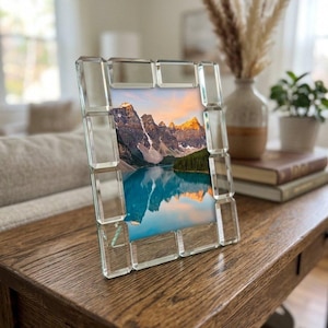 May include: A rectangular picture frame with a mirrored, beveled glass border. The frame contains a photograph of a mountain range with snow-capped peaks reflecting in a bright blue lake under a soft, warm-toned sky at sunrise or sunset.
