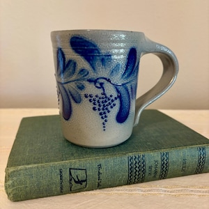 May include: A light gray stoneware mug with a textured surface, featuring hand-painted cobalt blue leaf and grape cluster motifs. The mug has a sturdy handle and a cylindrical body. The mug is resting on a green hardcover book titled Heidi.