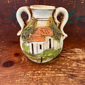 May include: A cream-colored ceramic vase with two handles, featuring a raised relief design of a house with an orange roof, green foliage, and painted details. The vessel has a rounded body and a wide opening, showcasing a textured finish.