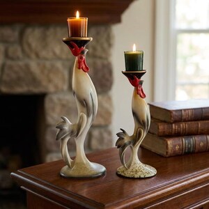 May include: Two rooster-shaped candle holders made of painted ceramic. The taller holder features a brown candle, while the shorter one holds a dark green candle. Both roosters have cream-colored bodies with gray accents and bright red combs.