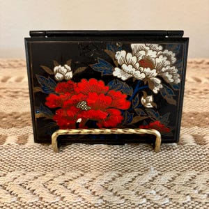 May include: A rectangular black compact case featuring a floral design with large red and white peony-like flowers, blue leaves, and gold accents. The case is hinged at the top and is displayed on a small, gold-toned metal wire stand.