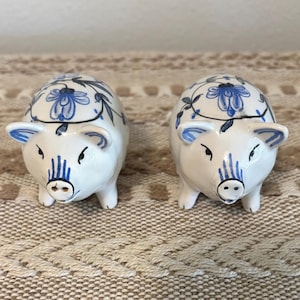 May include: A pair of white ceramic pig-shaped salt and pepper shakers featuring hand-painted blue floral patterns on the back, blue striped snouts, and blue-lined ears. Each pig has two small holes on the snout for dispensing seasoning.