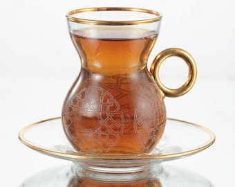May include: A clear glass tea cup with a curved waist and a gold rim, filled with amber tea. The cup features a circular gold handle and etched geometric patterns on the body, resting on a matching clear glass saucer with a gold-trimmed edge.