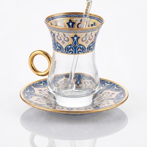May include: A clear glass teacup with a gold handle, on a matching saucer. The cup and saucer have a blue and gold ornate pattern. A small spoon sits in the cup. The set is designed for tea, with a decorative style.