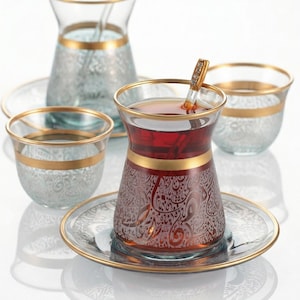 May include: A set of ornate glass teacups and saucers with gold trim. One cup is filled with tea and has a spoon. The other cups are empty. The glasses have a decorative etched pattern.