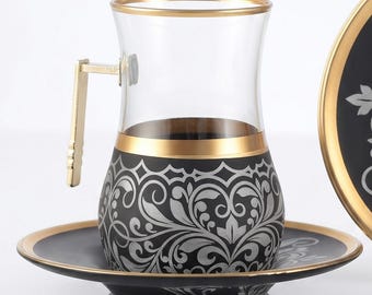 May include: A clear glass tea cup with a gold rim and a black base featuring a silver floral pattern, paired with a matching black saucer with a gold rim and silver floral details. The cup includes a gold-colored metal handle attached to the side.