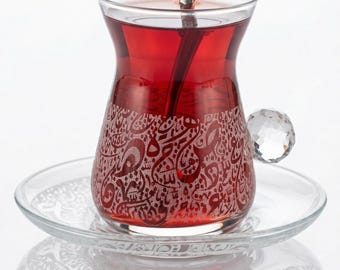 May include: A clear glass tea cup filled with red liquid, resting on a matching saucer. The cup features an etched design of Arabic calligraphy. A small silver spoon rests inside the cup. The handle is a clear, faceted crystal ball.