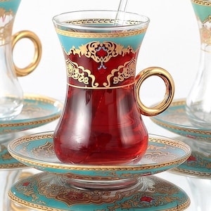May include: Clear glass teacups with ornate gold and teal designs, filled with red tea, sit on matching saucers. Each cup has a gold handle and a clear glass spoon. The set is ideal for serving tea.