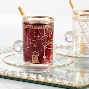 May include: Two glass teacups with gold rims and handles, filled with a red liquid. The cups are decorated with gold patterns and sit on clear saucers. Each cup has a gold-tipped stirrer. The cups are on a mirrored tray.
