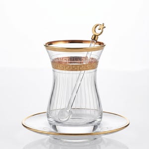 May include: Clear glass tea cup with gold trim and a matching saucer. A gold-coloured spoon with a crescent moon and star design rests inside the cup. The cup has a decorative band with a repeating pattern.