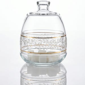 May include: A clear glass sugar bowl with a lid, featuring a gold band and a white leaf pattern. The bowl contains white sugar cubes. The design is elegant and suitable for serving sugar at a table setting.