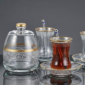 May include: A clear glass sugar bowl with a lid, accompanied by tea glasses on saucers, each featuring gold rims and white filigree patterns. A glass spoon with a blue and white evil eye bead at the top rests inside one of the tea glasses.