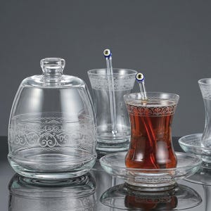 Op de afbeelding: Transparante glazen theeset met geëtste bloemmotieven. Inclusief een suikerpot met deksel, theeglazen, schoteltjes en roerstaafjes met blauwe oogvormige details. Eén glas is gevuld met thee.