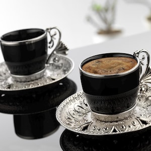 Pode incluir: Duas xícaras de café turco pretas e prateadas com seus pires. As xícaras têm uma borda prateada e os pires são adornados com intrincados desenhos prateados.