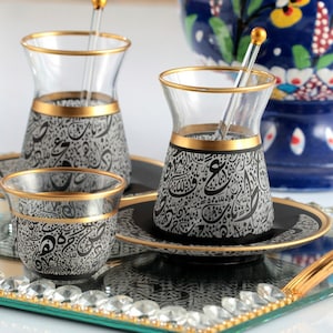 Pode incluir: Um conjunto de xícaras de chá e pires de vidro transparente com bordas douradas e um design de caligrafia preto e cinza. Cada xícara tem um pires correspondente e um misturador de vidro transparente com uma bolinha dourada.