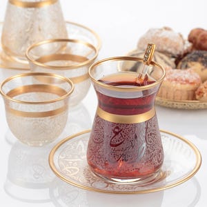 May include: Elegant glass tea set with gold trim. The set includes a teapot, teacups, and saucers, all adorned with intricate patterns. A teacup filled with tea sits on a saucer, with a decorative spoon inside. Baked goods are in the background.
