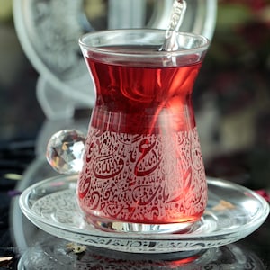 May include: A clear glass teacup with a red tea inside and intricate etched designs. The teacup has a diamond-shaped handle and sits on a matching saucer.